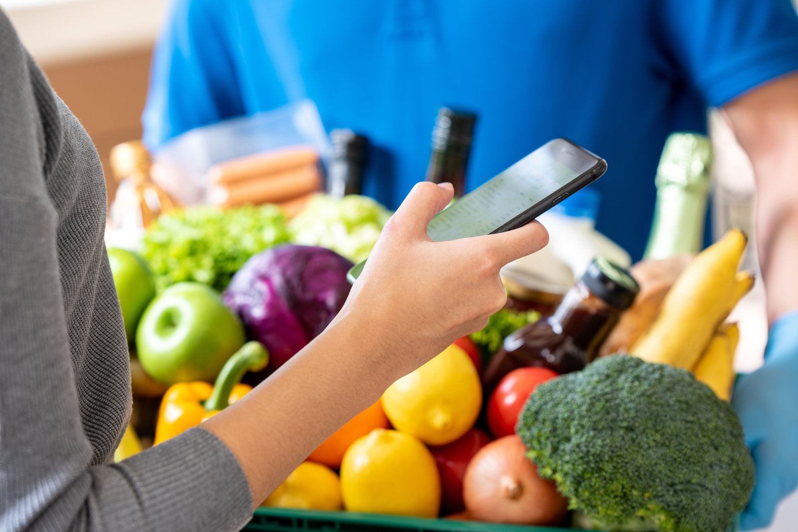 Holding phone and grocery basket full of colorful fruits and vegetables.